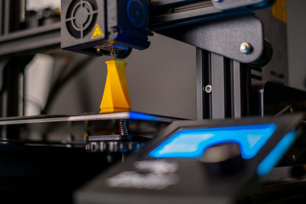 Close-up of a 3D printer in operation, with a yellow object being printed on the build plate and the printer's control panel visible.