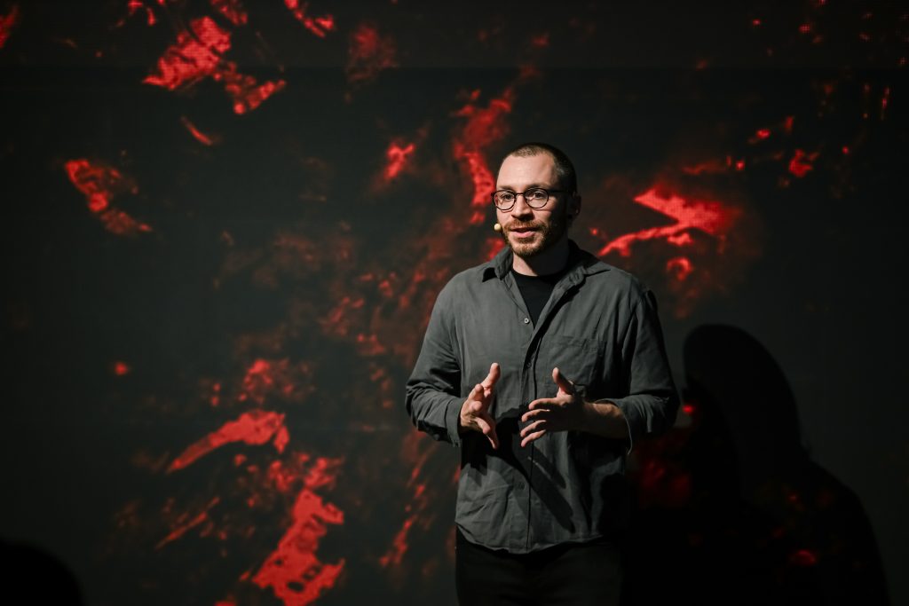 A speaker stands in front of a dark, abstract background with red lighting, gesturing while discussing a topic.