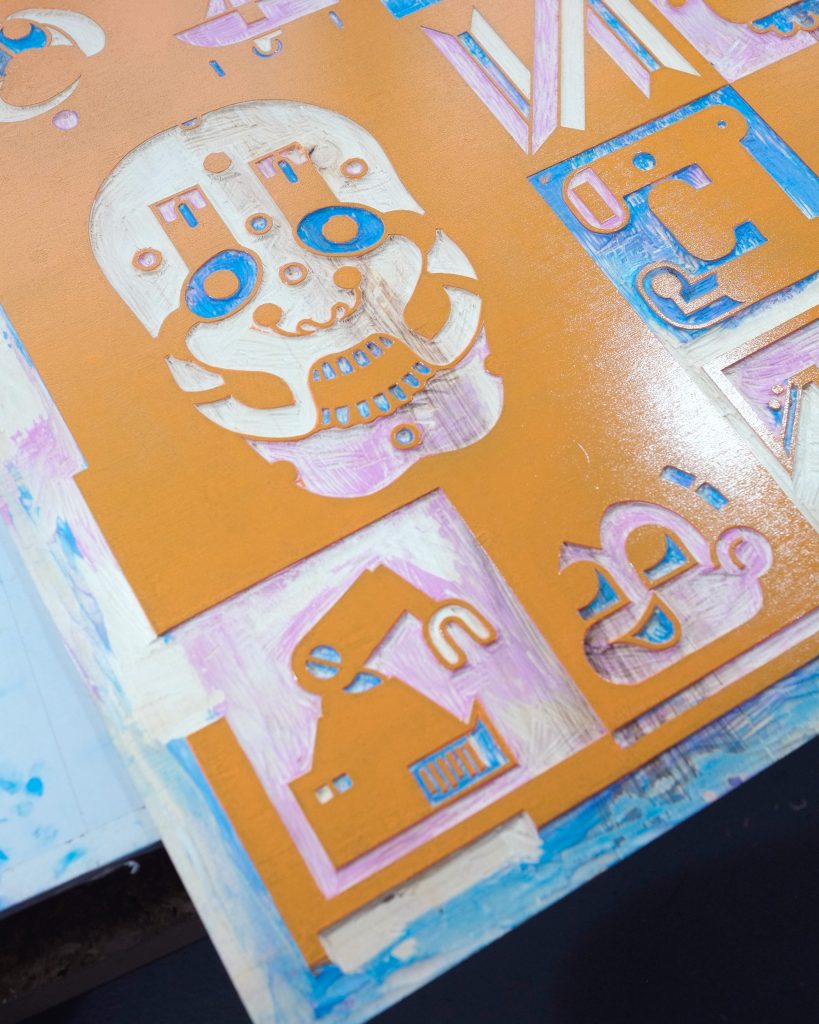 A close-up view of an intricate, orange printing plate featuring various carved designs, including a skull and abstract shapes, with blue and purple paint accents.