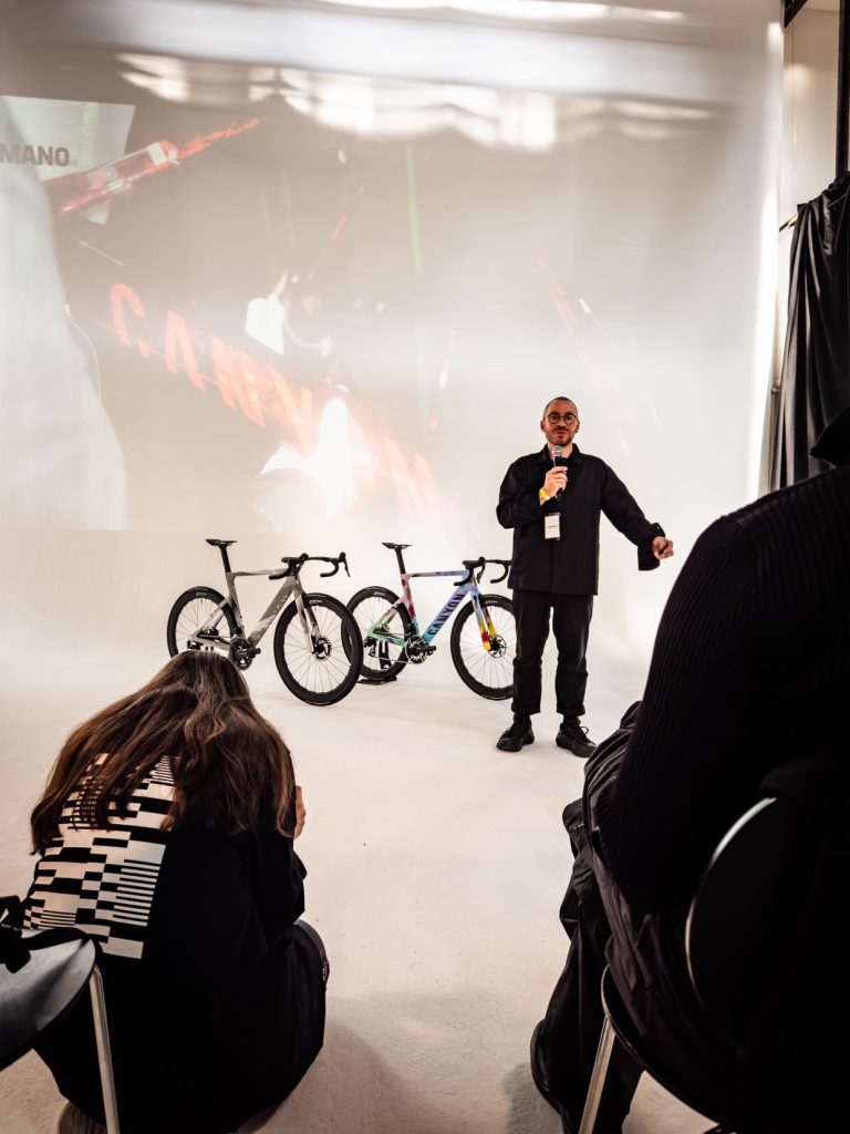 A speaker presents in front of two bicycles on display in a studio setting, while an audience member takes notes in the foreground.