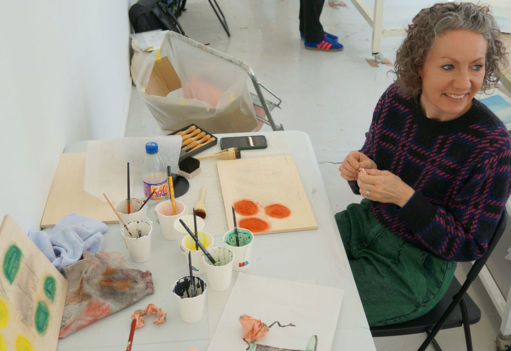 An artist sitting at a table filled with paint supplies, including brushes and paint containers, while smiling and working on a wooden canvas.