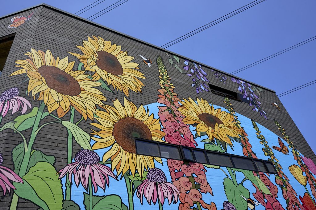 A colorful mural depicting large sunflowers, purple foxgloves, and various garden flowers on a building wall, against a clear blue sky.