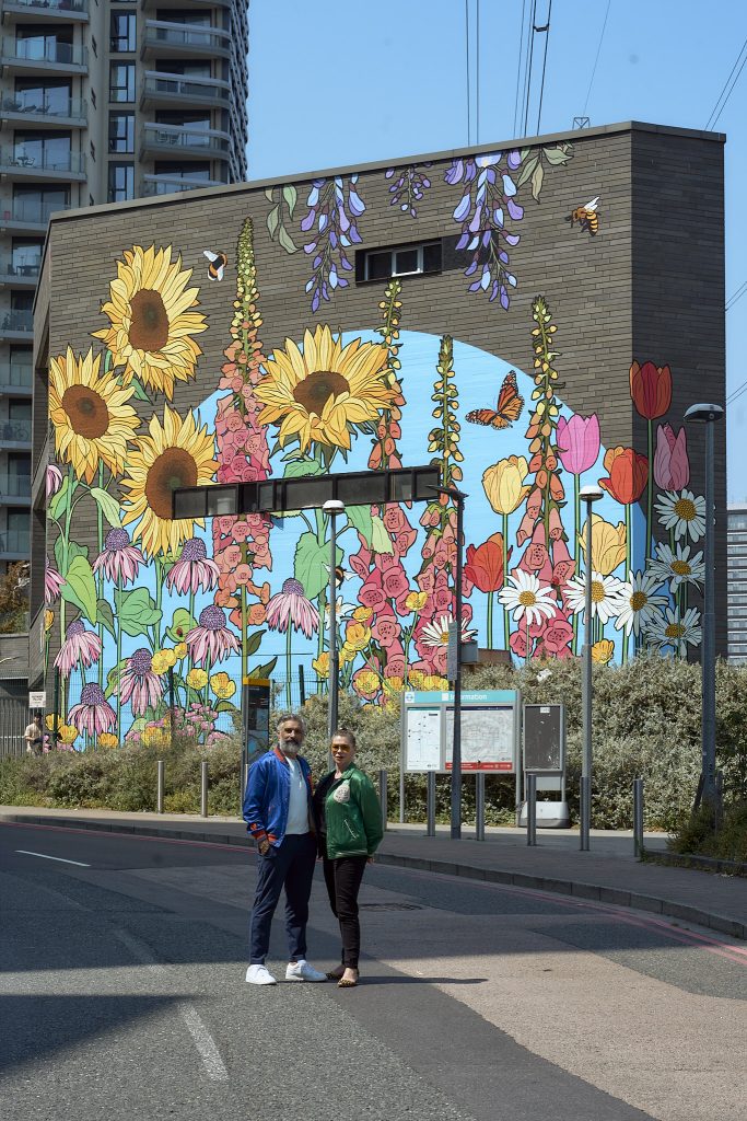 A colorful mural featuring sunflowers, flowers, and butterflies on a building wall, with two people standing in front on a street.