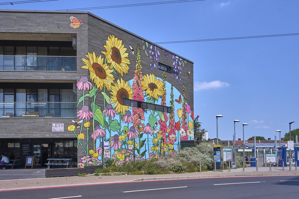 Colorful mural featuring sunflowers, wildflowers, and butterflies on the side of a modern building with a coffee shop and outdoor seating area.