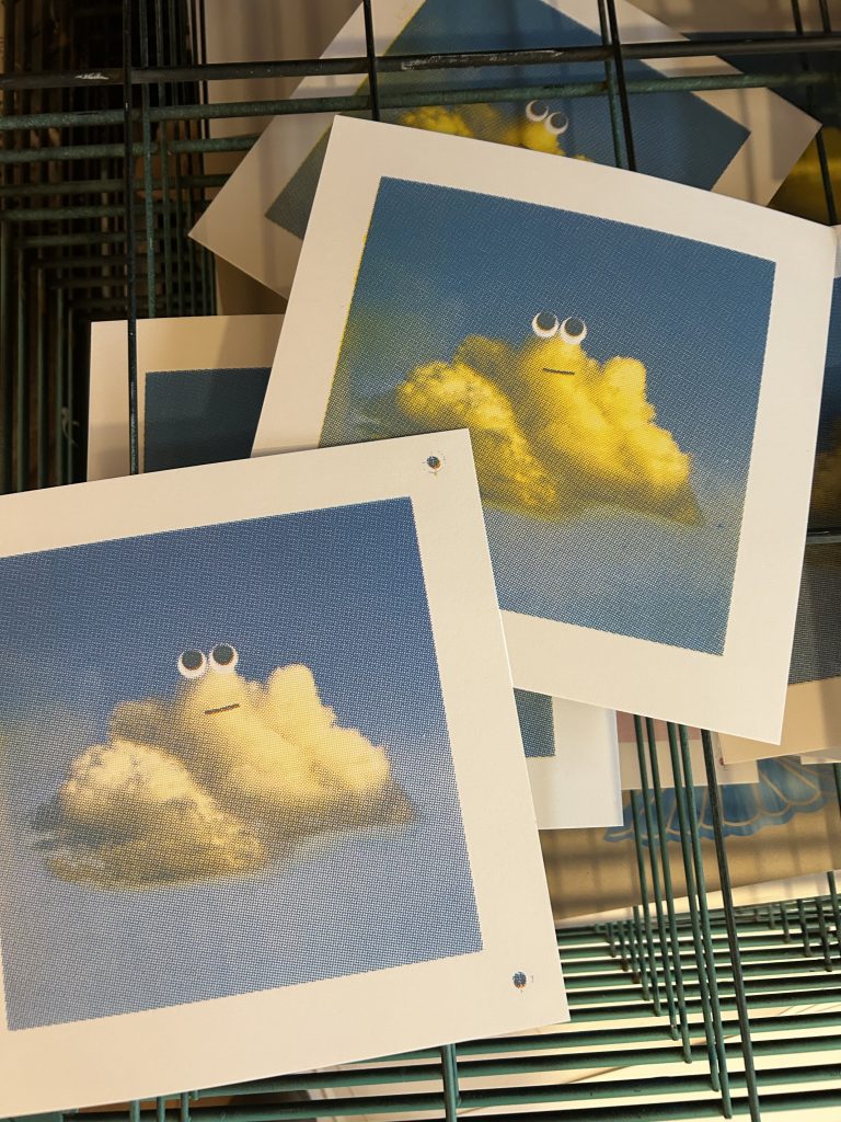 Art prints of fluffy clouds with googly eyes and simple expressions displayed on a wire rack.