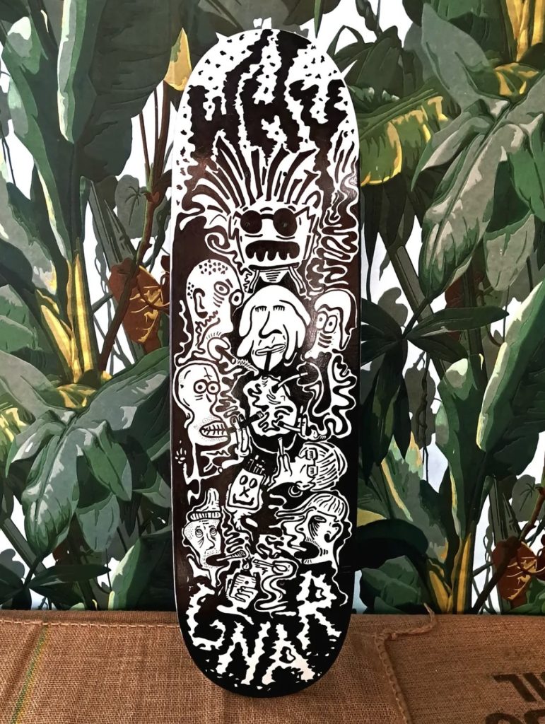 A skateboard deck featuring bold, cartoonish illustrations of various whimsical characters, including faces with exaggerated features and playful expressions. The words 'WHY' and 'GNAR' are prominently displayed in a stylized font. The backdrop consists of lush green foliage.