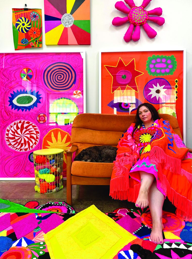 A woman sits on an orange couch, wearing a colorful, fringed blanket. She is surrounded by vibrant artwork featuring bold patterns and bright colors, with a small dog resting on the couch. A colorful rug and a basket of decorative yarns are in the foreground.
