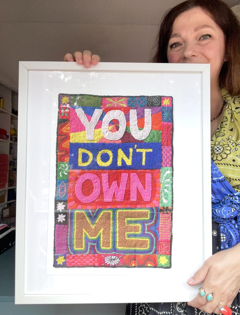 A woman holding a colorful framed textile artwork that says 'YOU DON'T OWN ME' in bold letters.
