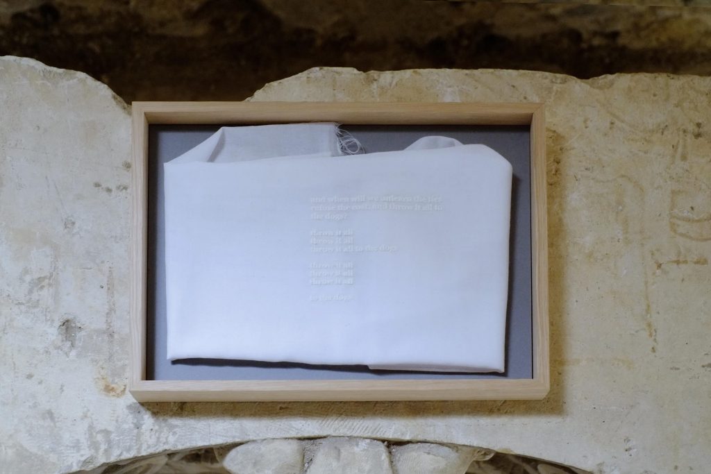 A framed piece of white fabric displayed against a textured stone wall, with partially visible text printed on it, surrounded by a light wooden frame.