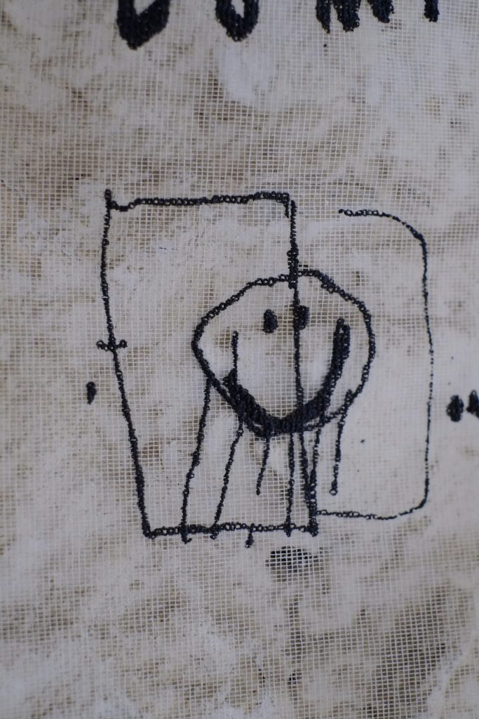 A close-up of a fabric with a simple black sketch of a smiling face surrounded by a square, with faint text above and below.