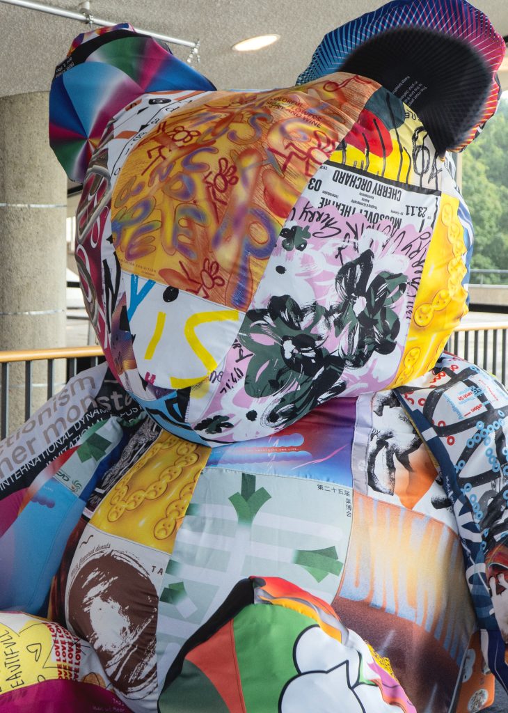 A large, colorful bear sculpture composed of various fabric patches featuring vibrant designs, text, and patterns.