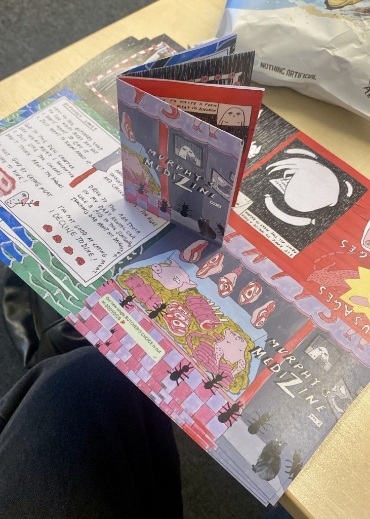 A colorful zine titled 'Murphy's Medizine' is opened on a table, displaying illustrated pages with vibrant designs, including cartoon-like characters and various food illustrations, such as meats and vegetables. Notes in handwriting are visible on some pages.