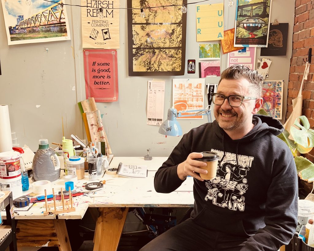 A smiling person sitting at a cluttered art studio desk, holding a coffee cup. The background features various colorful posters and art supplies scattered around.