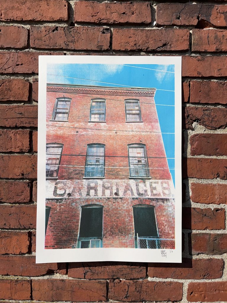 A photograph of an old brick building with the word 'CARRIAGES' painted on it, displayed against a brick wall.