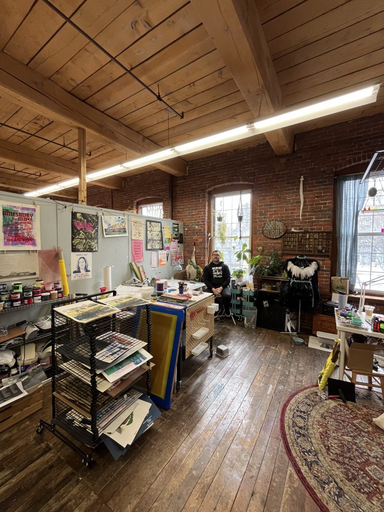 An artist's studio featuring a wooden floor, exposed brick walls, and large windows. Various art supplies are organized on tables, including paintings and printmaking materials, with a person seated in the background.