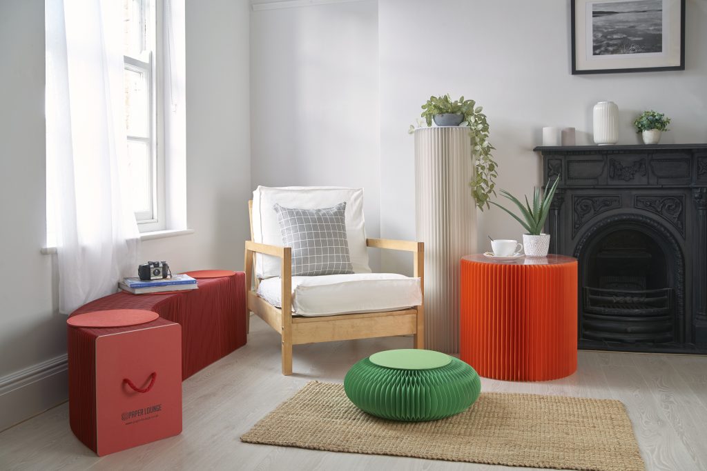 A modern living room featuring a wooden chair with white cushions, colorful pleated furniture including a red table and green pouf, a decorative fireplace, and plants. Natural light filters through sheer curtains.