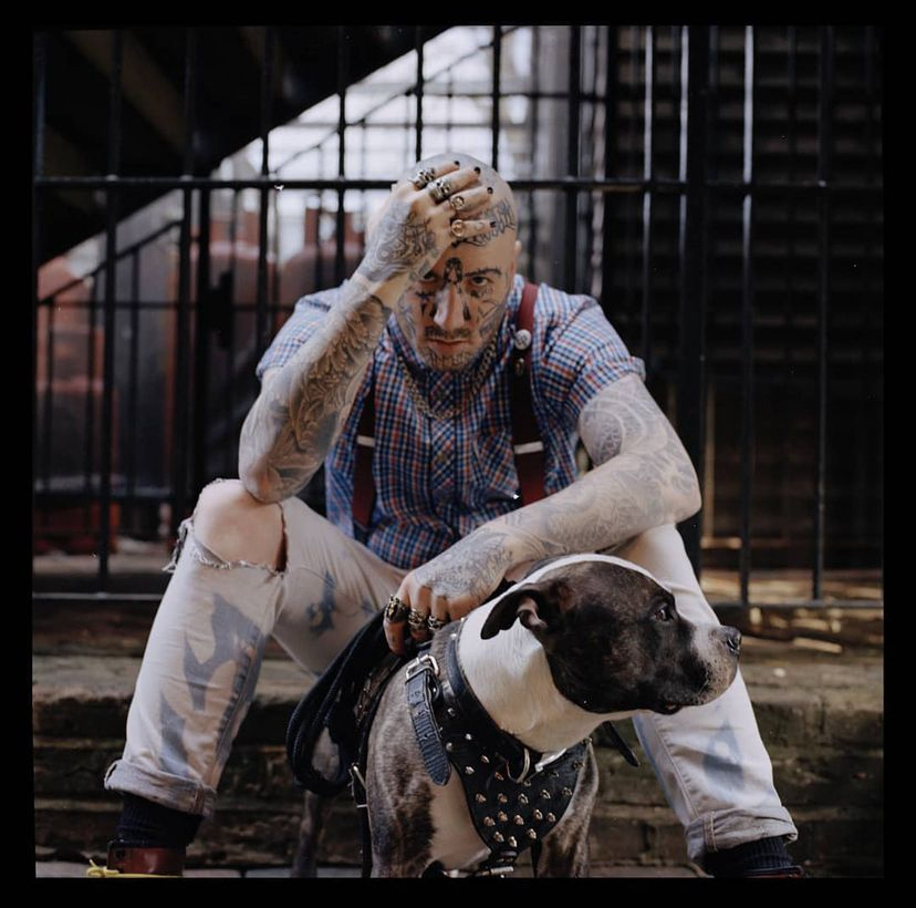 A tattooed individual sitting on steps, wearing a checkered shirt and suspenders, with one hand on their head. A leashed dog sits beside them, looking off to the side.