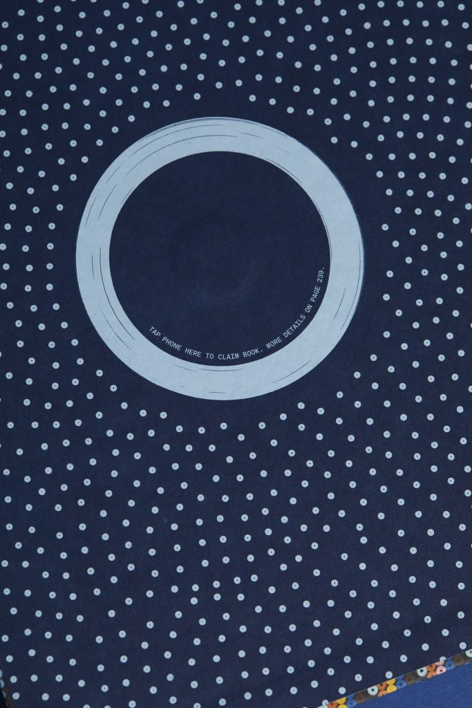 A dark blue background with small white polka dots, featuring a circular design in the center with text that says 'TAP PHONE HERE TO CLAIM BOOK. MORE DETAILS ON PAGE 239.'