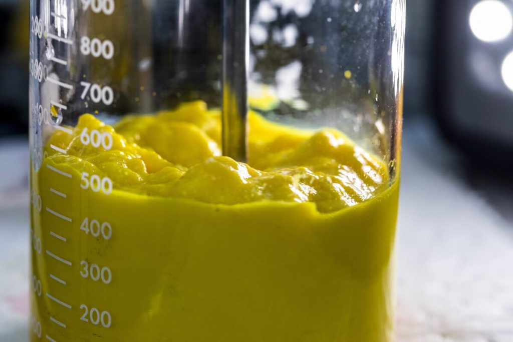 A close-up of a beaker filled with bright yellow liquid, showing a mixing tool submerged in it.