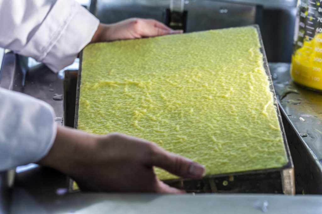 A person holding a tray filled with a textured, yellow substance in a kitchen or food processing environment.
