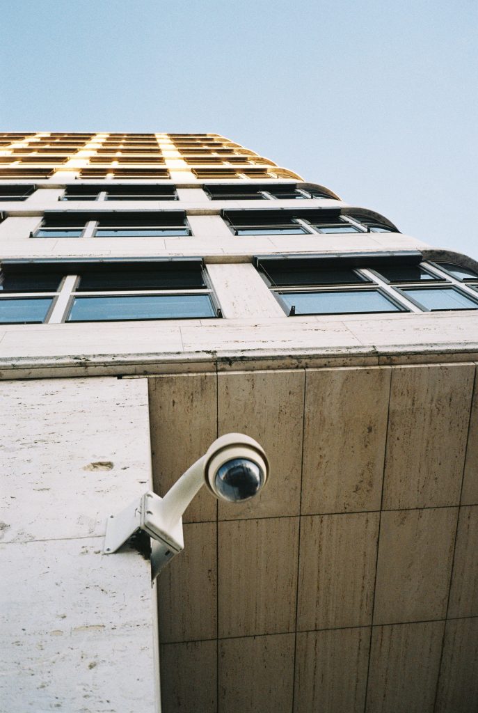 Looking up at a tall building with a surveillance camera mounted on the corner, against a clear blue sky.