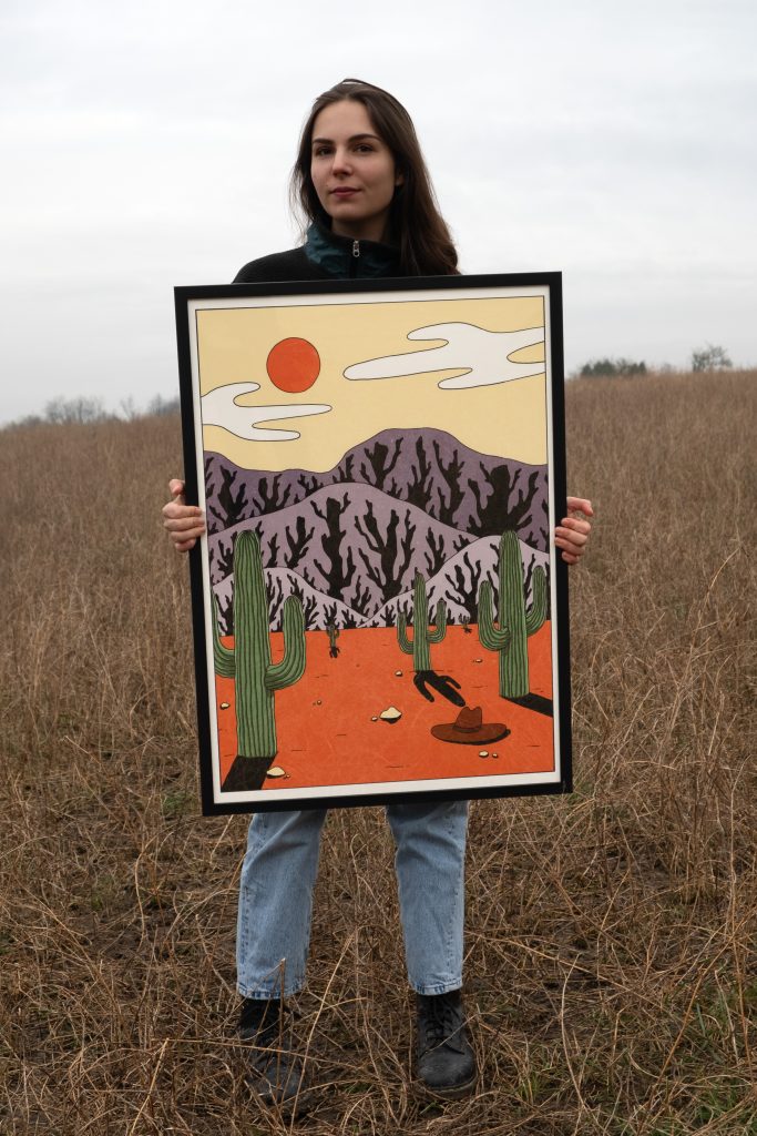 A woman standing in a field holds a framed illustration depicting cacti and mountains under a bright orange sun.