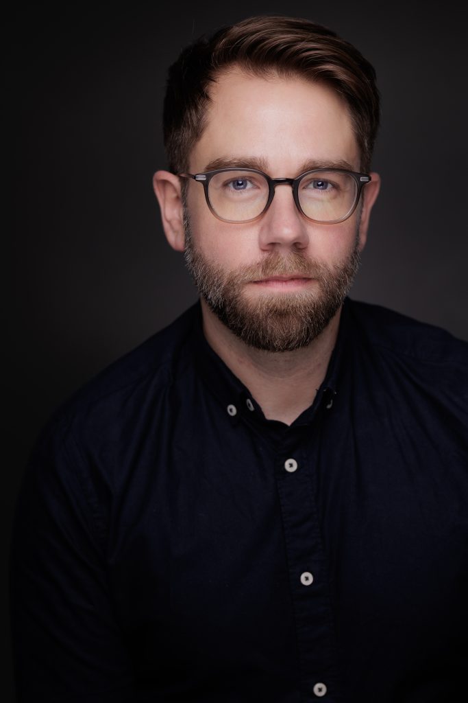 A close-up portrait of a man with short, dark hair and a beard, wearing glasses and a black button-up shirt, against a dark background.