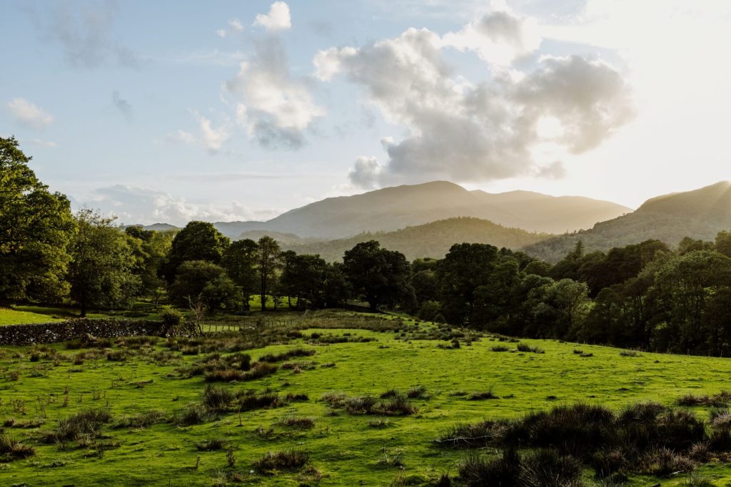 A scenic landscape featuring green fields, trees, and mountains under a cloudy sky during sunset.