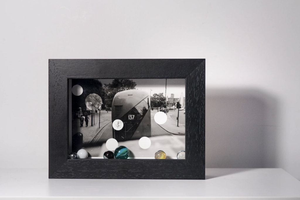 A black-framed shadow box featuring a black-and-white photograph of a street scene with a tram, enhanced by colorful marbles in the foreground.