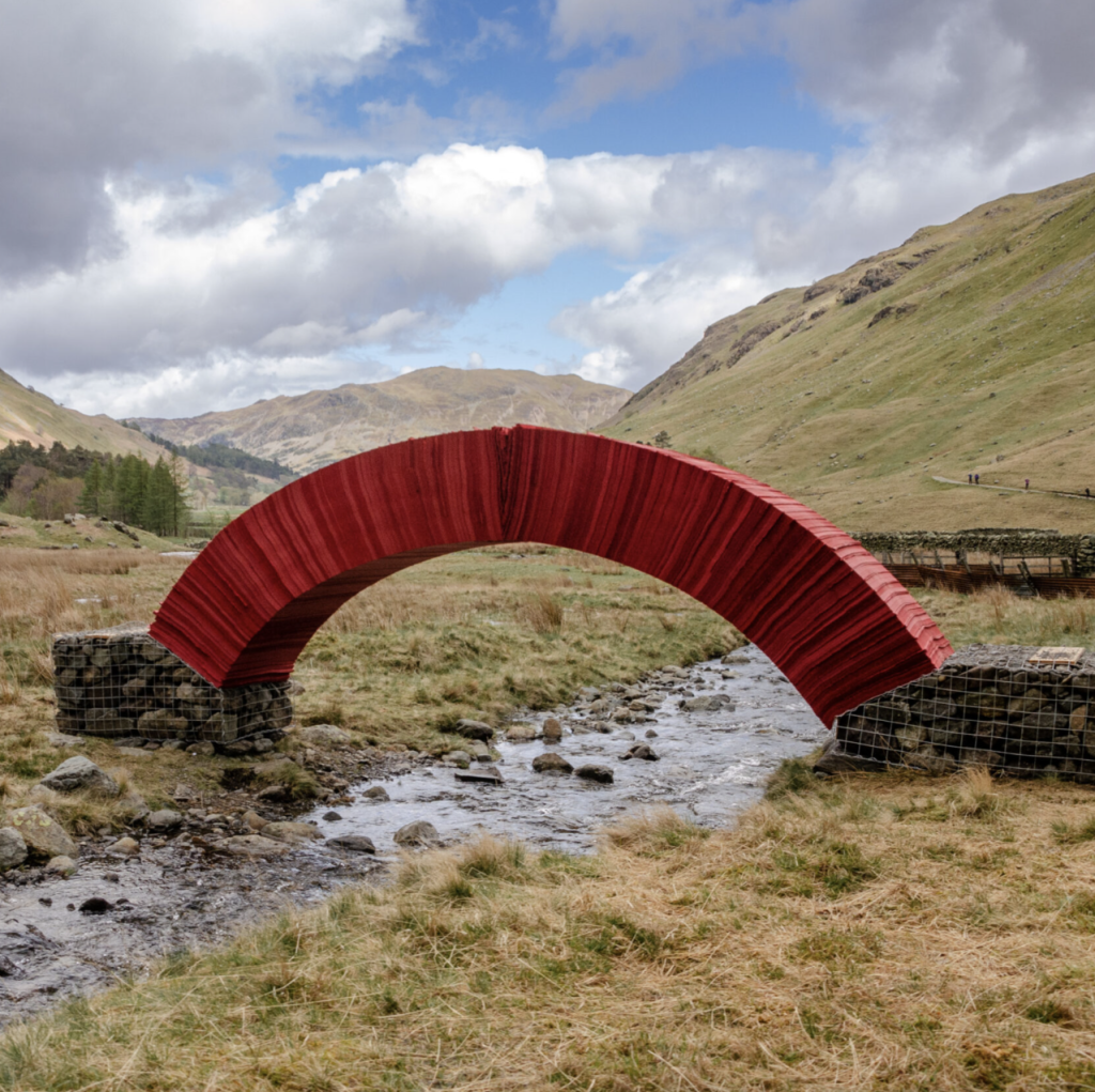A bright red arched bridge over a small stream, surrounded by grassy fields and hills under a cloudy sky.