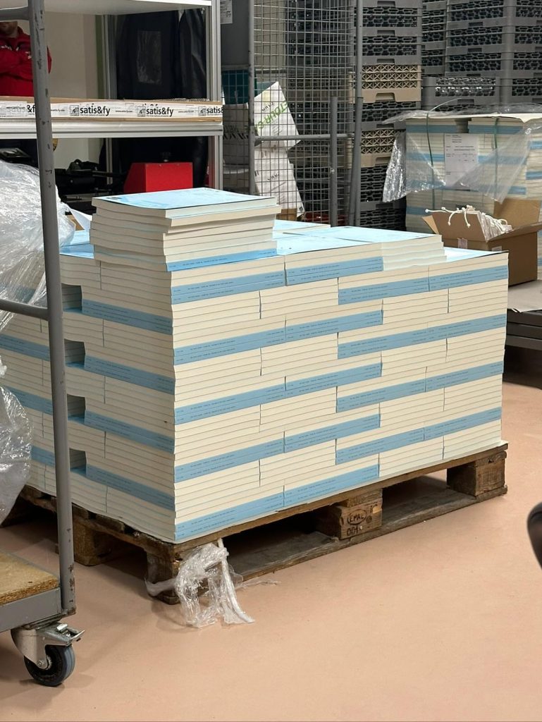 A stack of neatly arranged books with blue edges resting on a wooden pallet in a warehouse setting.