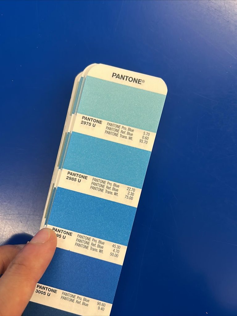 A hand holding a Pantone color guide displaying various shades of blue, including Pantone 2975 U, 2985 U, 295 U, and 3005 U, on a blue table.