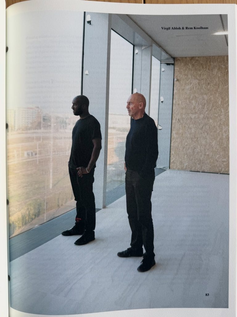 Two men standing inside a modern building, looking out of large windows. The first man, wearing a black t-shirt, is facing slightly to the side, while the second man, with a shaved head, is standing nearby in profile. The background features a view of the outdoors.