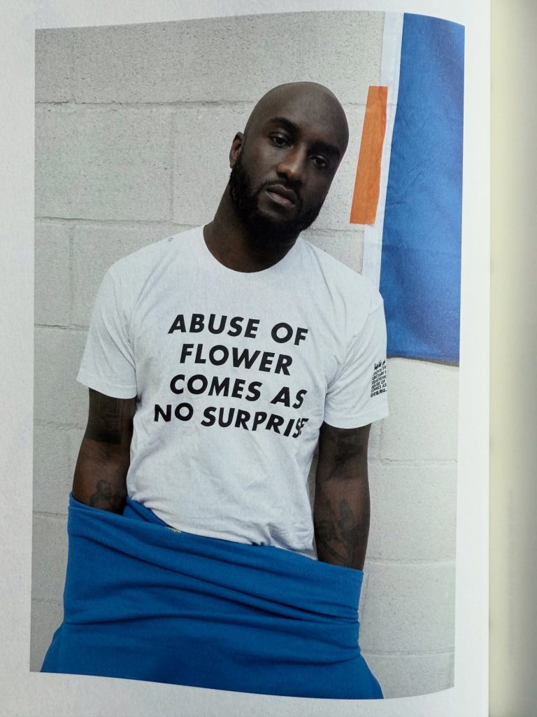 A person wearing a white t-shirt with the text 'ABUSE OF FLOWER COMES AS NO SURPRISE' stands against a gray wall, dressed in blue pants.