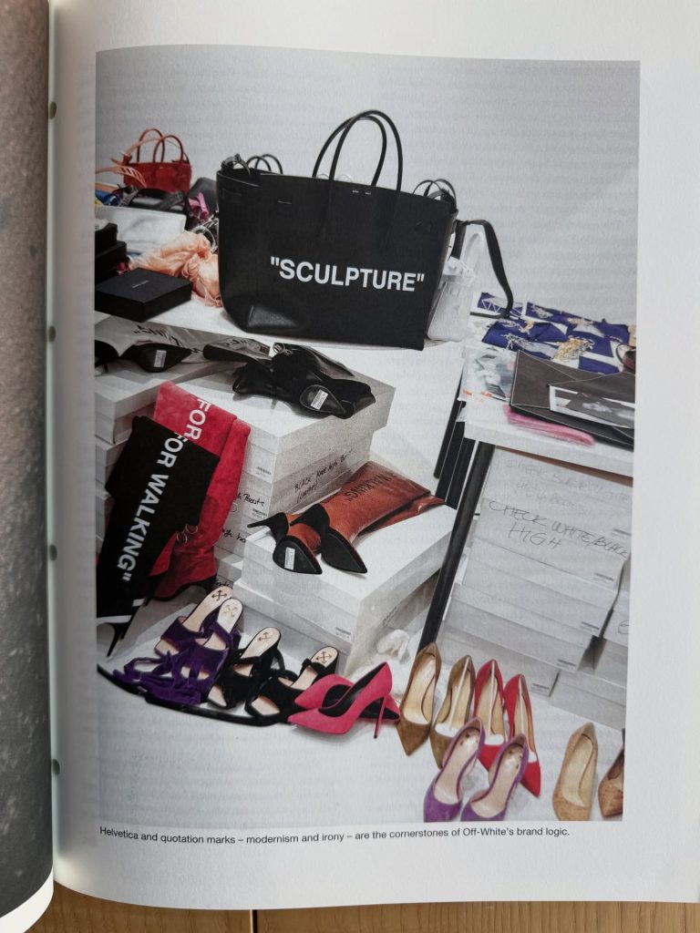 An assortment of shoes displayed on a table, featuring high heels in various colors, alongside a large black bag with 'SCULPTURE' printed on it, and scattered fashion items and boxes.