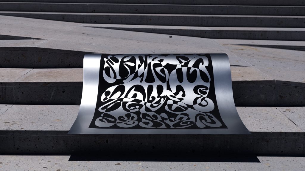 A metallic sheet displaying intricate, stylized lettering, resting on concrete steps. The design features black text that casts a shadow on the surface below.