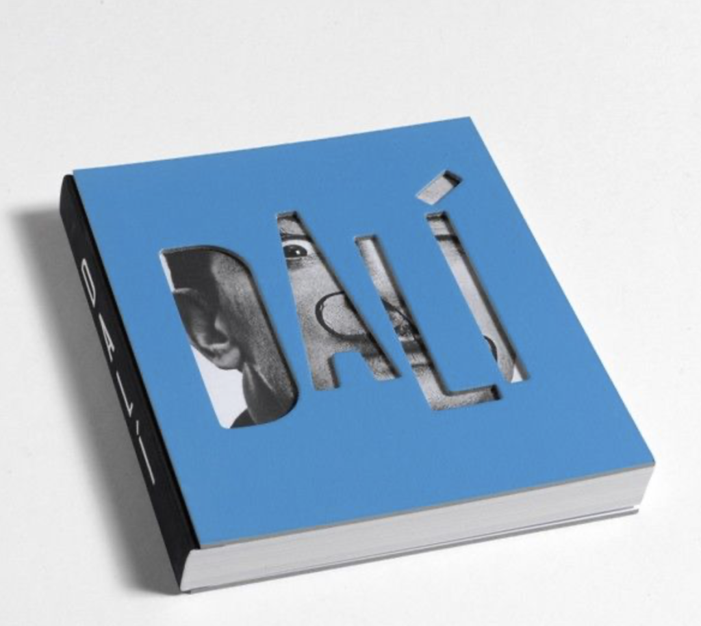 Close-up of a blue book cover featuring cut-out letters spelling 'DALÍ', revealing a black and white image beneath.