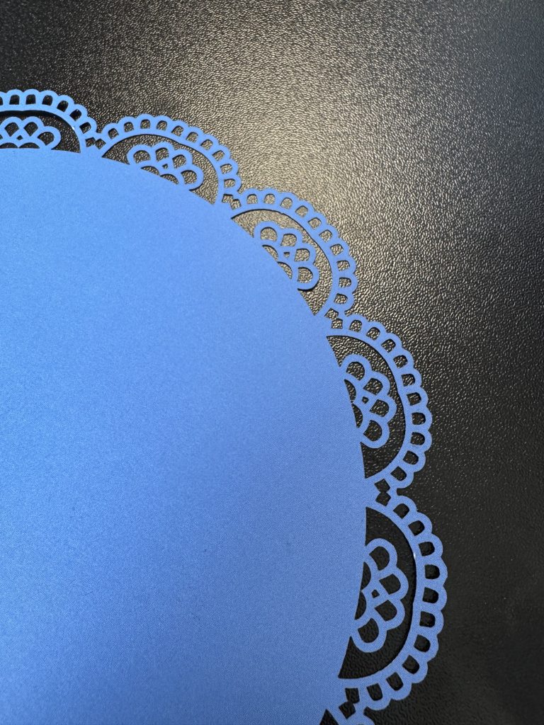 Close-up of a blue round paper doily with intricate cut-out designs, positioned on a black surface.